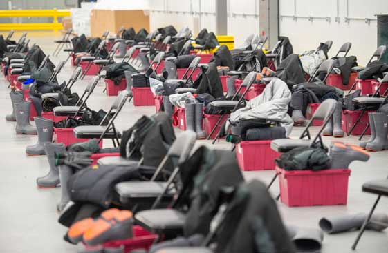 Folding chairs set up in rows in a large room with red plastic boxes and rubber boots and heavy jackets next to them.<br>  