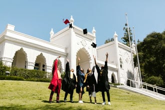 A group of graduates in graduation gowns stand on a lawn and throw their caps into the air.