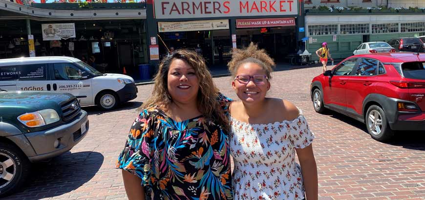 Two women smiling at the camera standing in front of a farmer's market