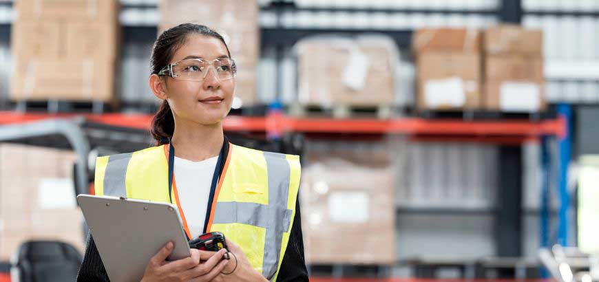 A female warehouse worker wearing safety glasses and a safety vest stands inside a facility holding a clipboard and scanner